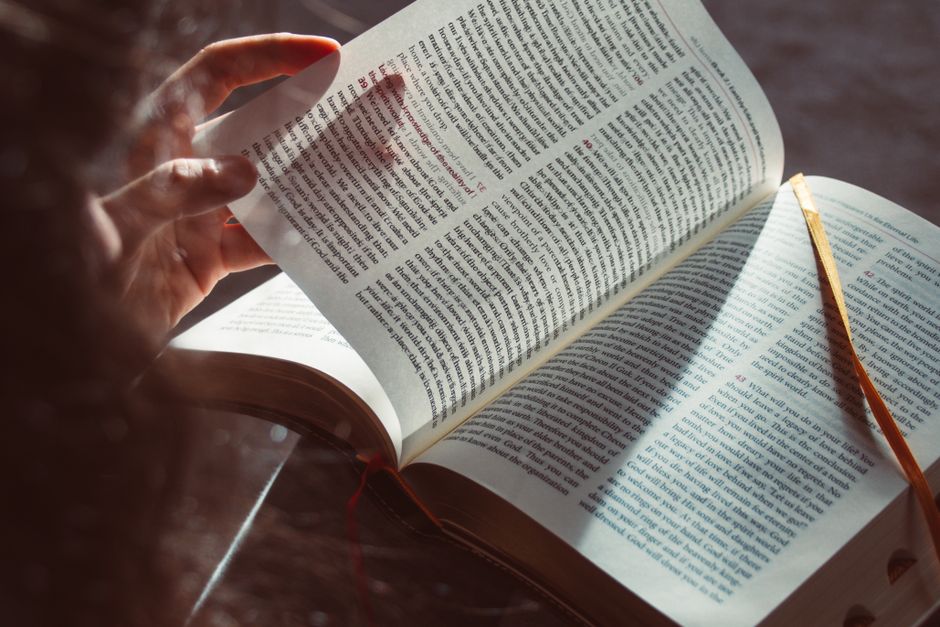 A hand gently turning a page of an open book in sunlight, creating a warm, tranquil reading scene.